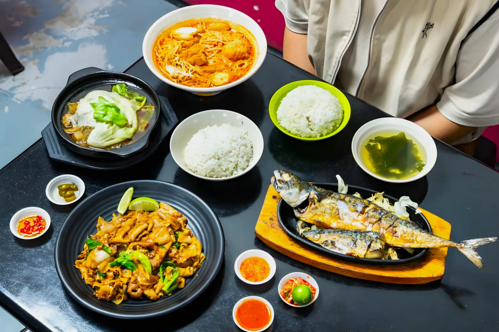 A colorful array of Asian dishes on a black table, featuring grilled fish, noodles, rice, soup, and sides. Warm tones convey a cozy dining experience.