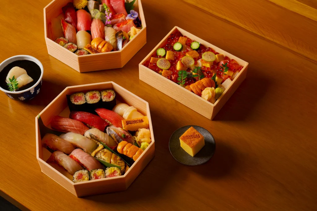 Assorted sushi and sashimi in wooden boxes on a table, featuring colorful nigiri, rolls, and garnishes. A cup of broth and a piece of cake accompany the meal.