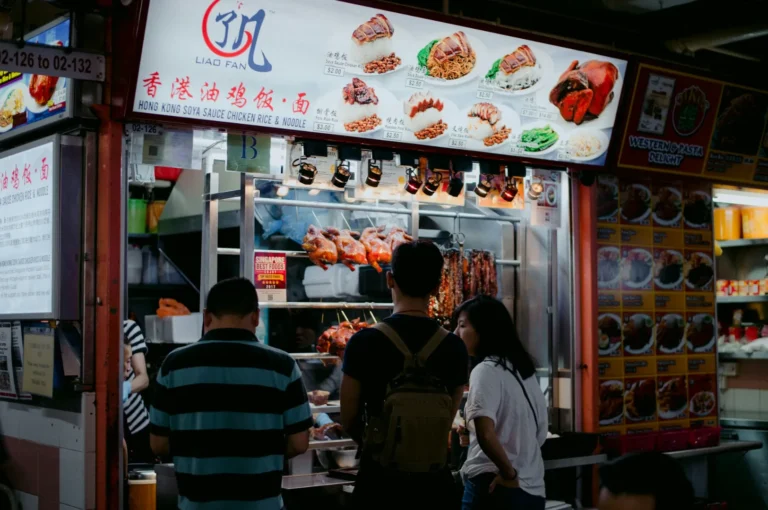 People stand in front of a bustling food stall at a market, with roasted meats on display and a menu signage featuring various Asian dishes. The atmosphere is lively and inviting.