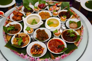 A round tray filled with various colorful Malaysian dishes in small bowls, including curries and soups, garnished with herbs on banana leaves.
