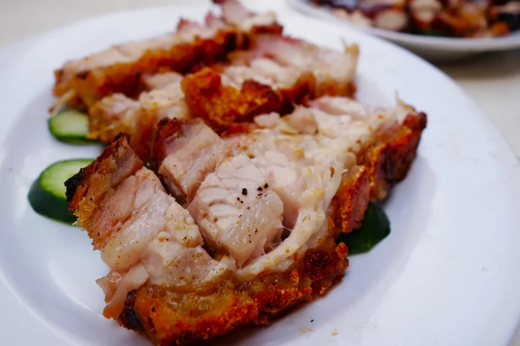 Close-up of crispy roasted pork belly slices on a white plate, placed over cucumber slices. The crackling is golden brown and inviting.