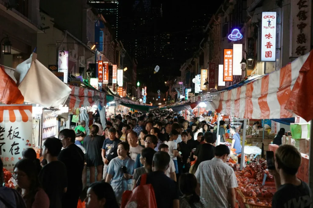 Bustling night market with a dense crowd browsing stalls under striped canopies. Bright, colorful signs illuminate the lively atmosphere.