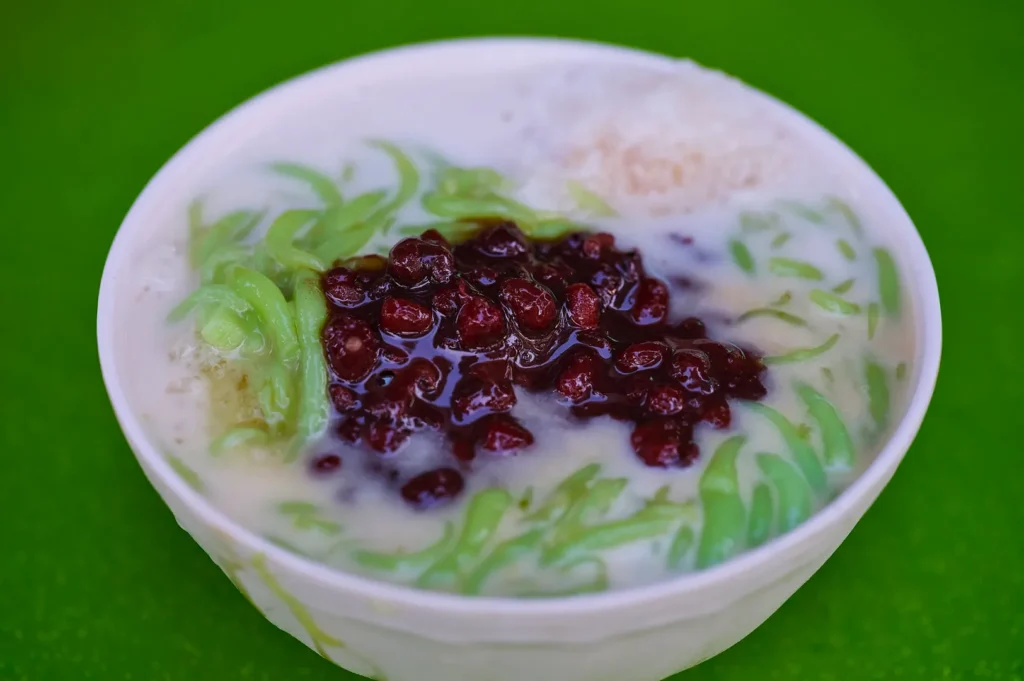 A bowl of cendol on a green background, featuring green rice flour jelly, red beans, coconut milk, and shaved ice, creating a refreshing dessert.