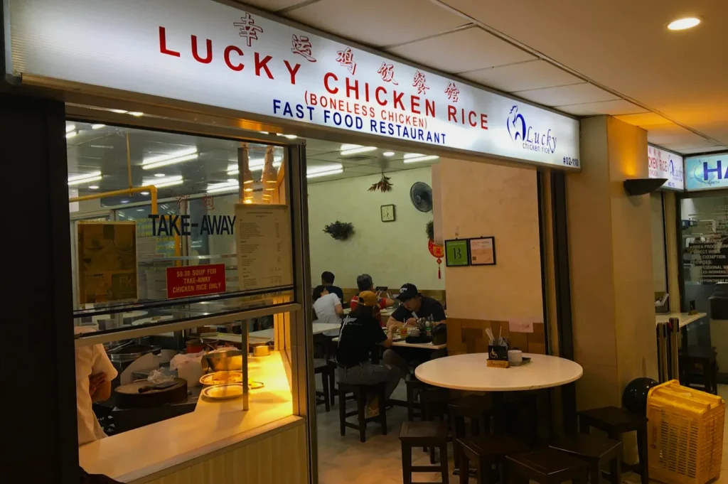 Restaurant with a sign reading "Lucky Chicken Rice," featuring a take-away counter and dining area. People are seated inside. Casual and cozy ambiance.