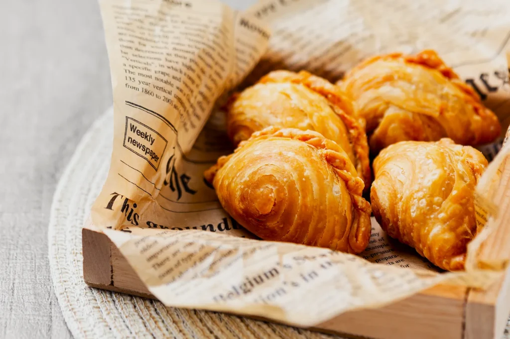Golden-brown pastries with flaky layers are nestled in a wooden tray lined with vintage-style newspaper. The scene is warm and inviting.