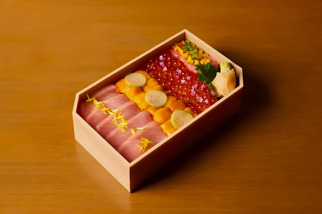 A wooden bento box on a wooden table features neatly arranged sushi ingredients: sliced fish, bright orange uni, red salmon roe, and wasabi, garnished with herbs.