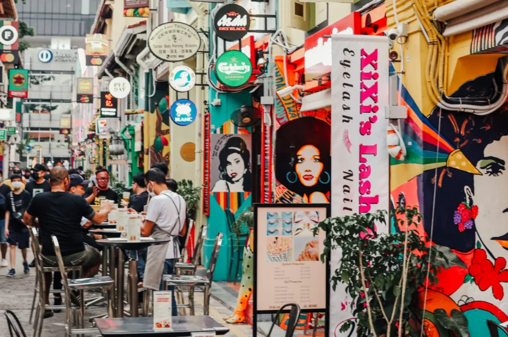 A vibrant, bustling street scene with colorful murals and diverse signage. People sit at outdoor tables, creating a lively, energetic atmosphere.