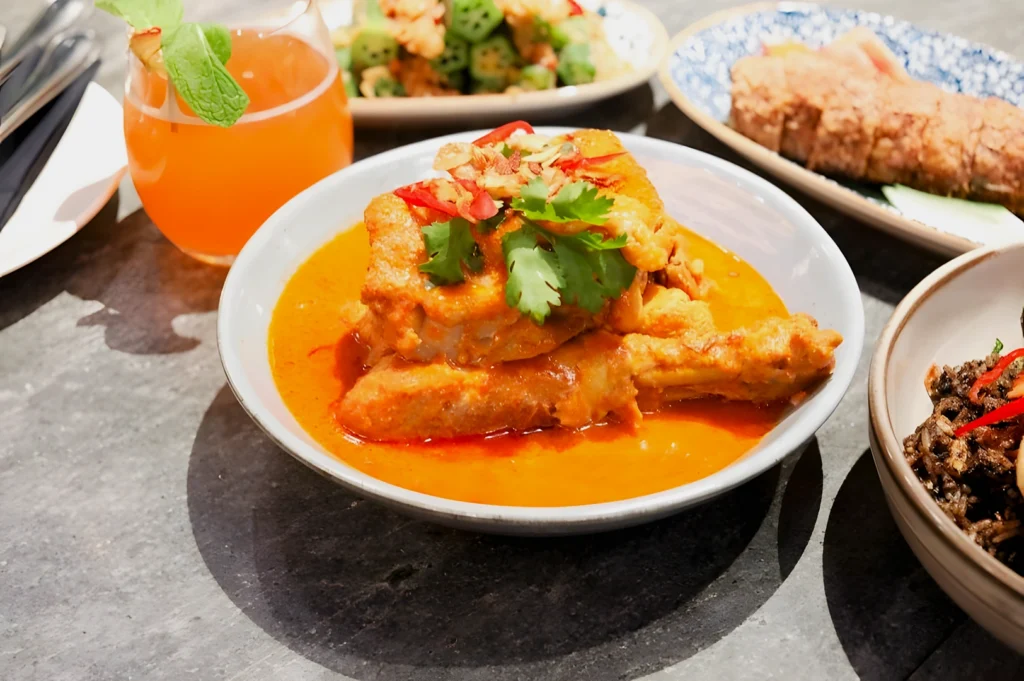 A bowl of vibrant red curry with chicken topped with cilantro and red chilies. Nearby are a glass of orange juice and other Asian dishes on a gray table.