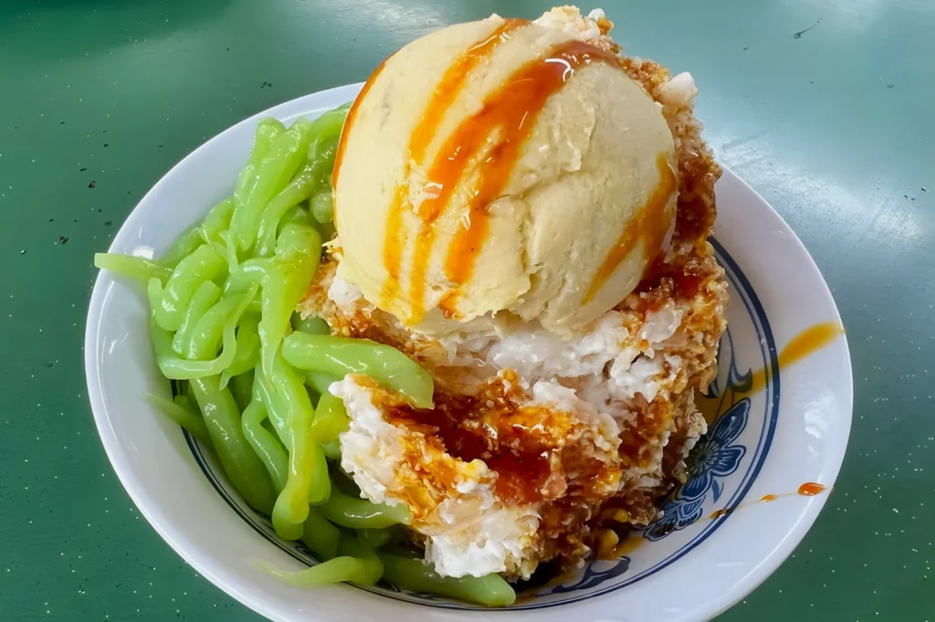 A bowl of traditional Malaysian dessert Cendol featuring a generous scoop of vanilla ice cream drizzled with syrup, on shaved ice and green pandan noodles.