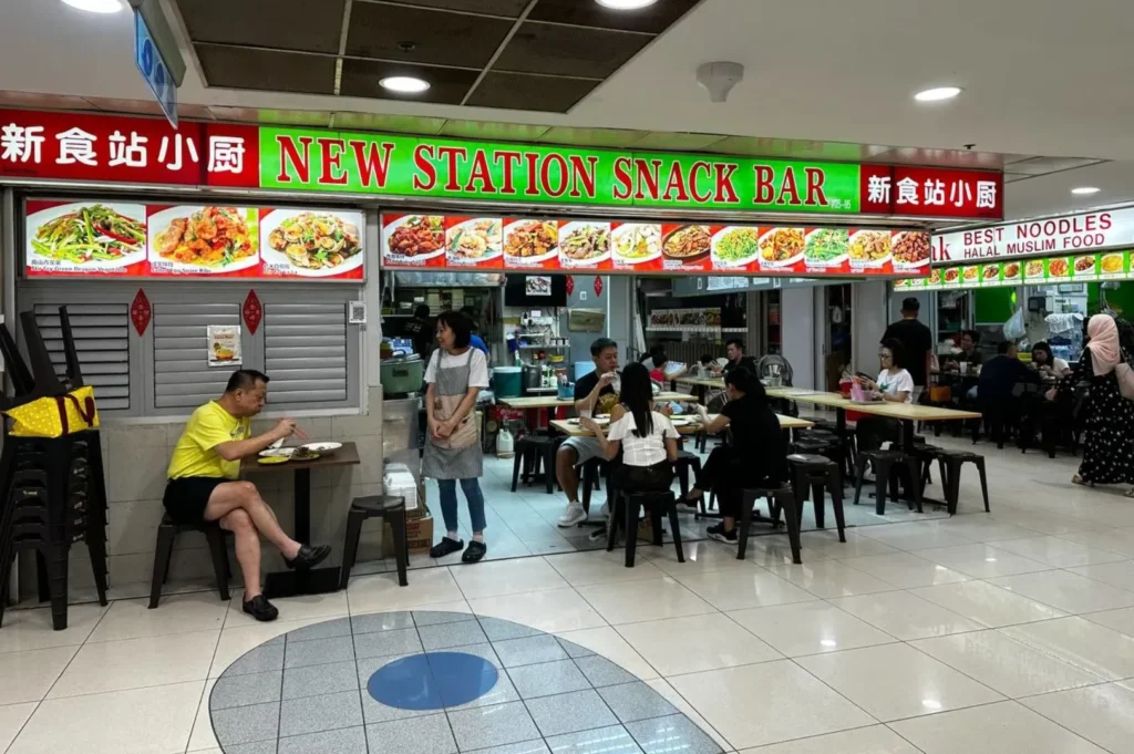 Hawker center scene showing "New Station Snack Bar," with patrons enjoying meals at tables. Bright signage showcases food photos, evoking a bustling, casual atmosphere.