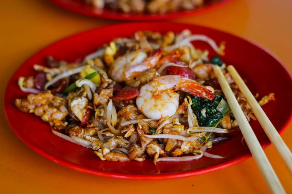 A vibrant red plate holds a serving of char kway teow with prawns, bean sprouts, and vegetables, garnished with chopsticks on a warm-toned table.