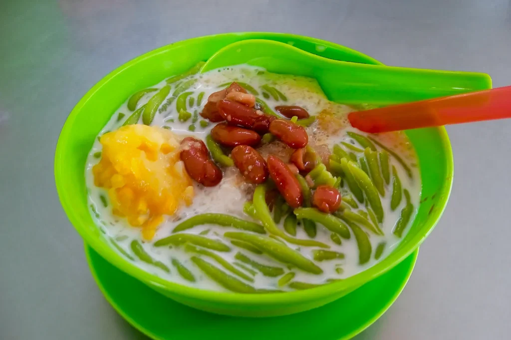 A vibrant green bowl of cendol sits on a matching saucer, containing green jelly noodles, red beans, corn, and coconut milk, evoking a refreshing vibe.