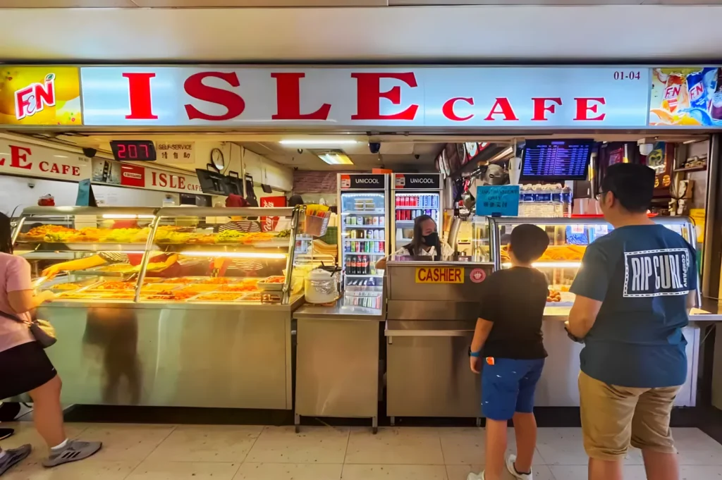 Brightly lit food stall named "ISLE Cafe" with a display of dishes in metal trays. A person serves customers while two people wait at the counter. Casual and busy atmosphere.