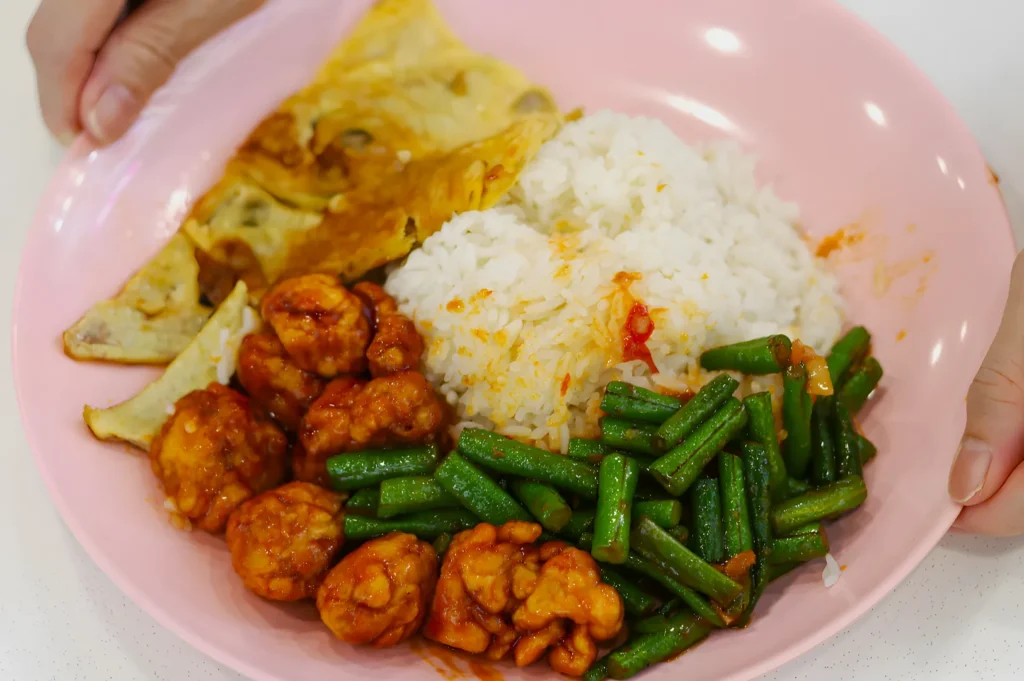 A pink plate holds a meal of white rice, spicy fried chicken, green beans, and a slice of omelette. A hand is supporting the plate.