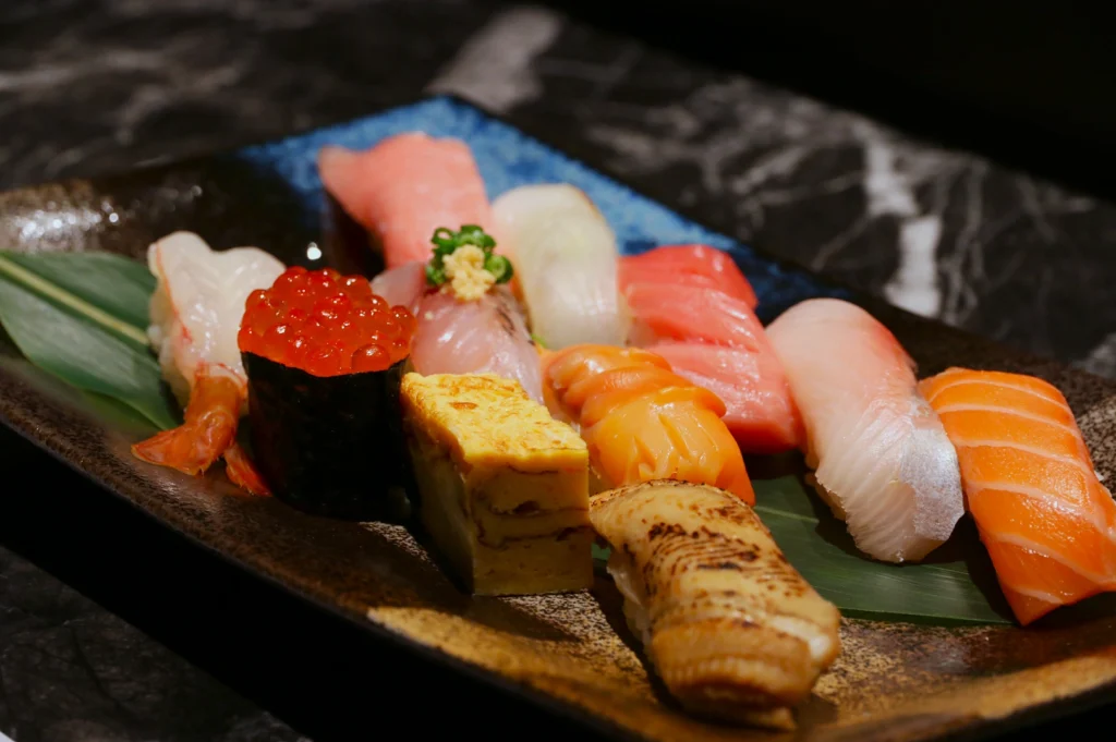 A tray of assorted sushi on a dark stone surface, featuring nigiri, sashimi, tamago, and ikura. Vibrant colors and textures create a fresh, elegant display.