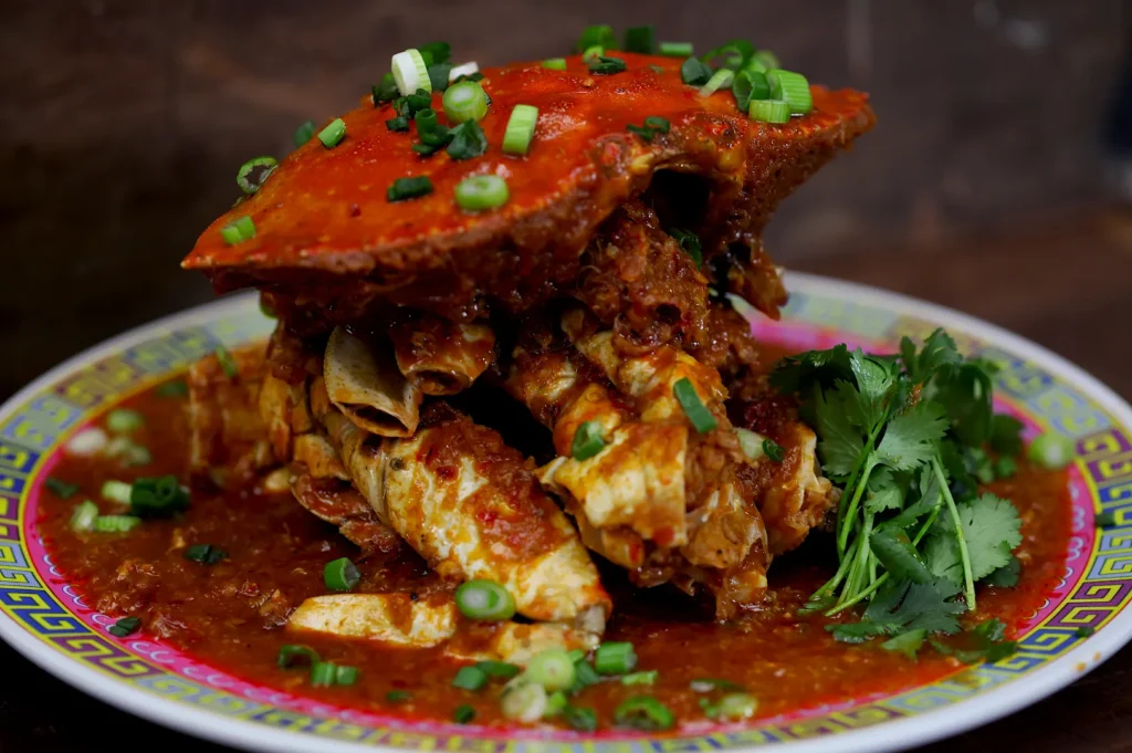 Vibrant dish of chili crab with a rich red sauce, topped with green onions. Garnished with cilantro on a colorful plate, evoking a spicy, savory aroma.