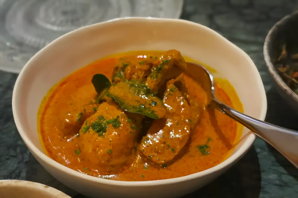 A white bowl filled with vibrant orange curry, garnished with green herbs. Visible chicken pieces and a spoon convey a savory, inviting meal.