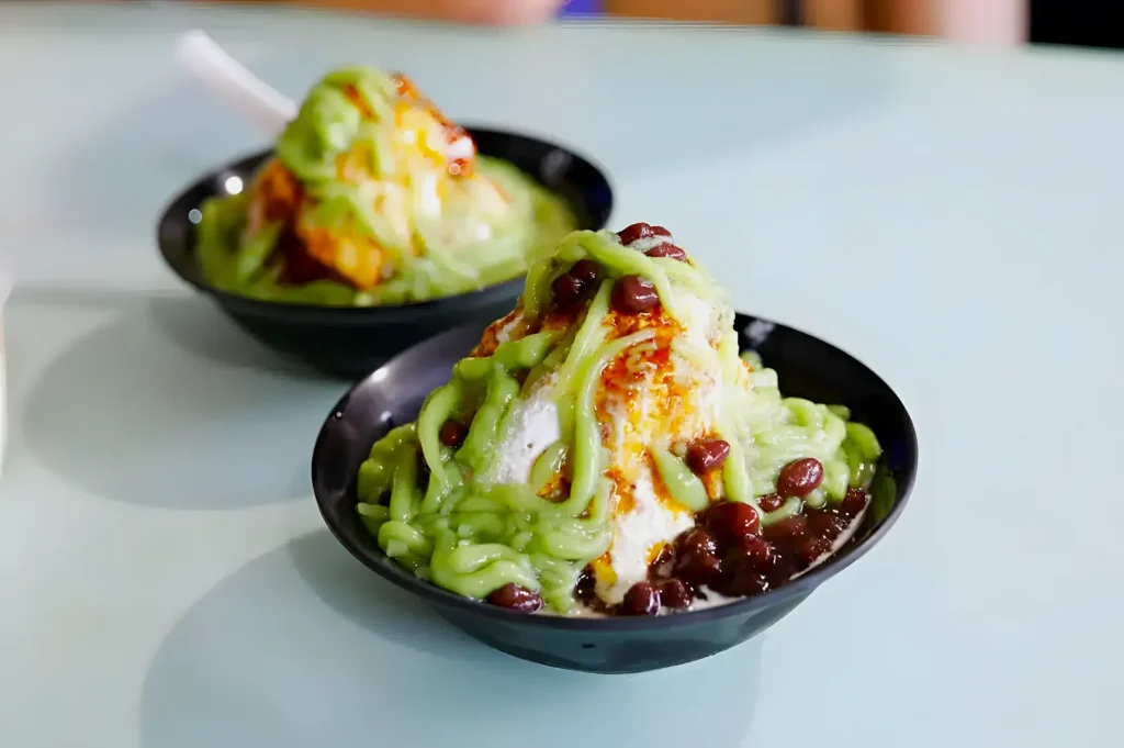 Two bowls of shaved ice topped with green pandan sauce and red beans, on a light blue table, creating a refreshing and inviting dessert scene.