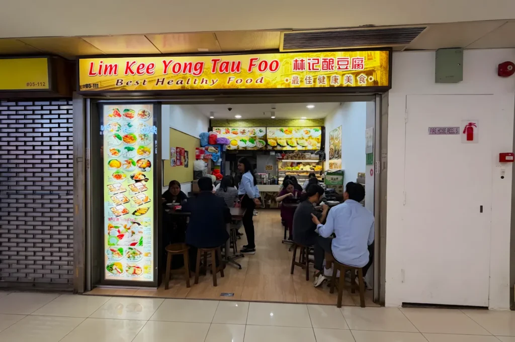 A small, brightly lit eatery named "Lim Kee Yong Tau Foo" with a glowing menu display on the left. Patrons sit inside at wooden tables, enjoying meals.