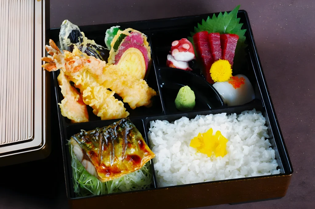 A bento box with rice topped with yellow garnish, tempura shrimp, mixed vegetables, grilled fish, and sashimi with wasabi. Colorful and neatly arranged.