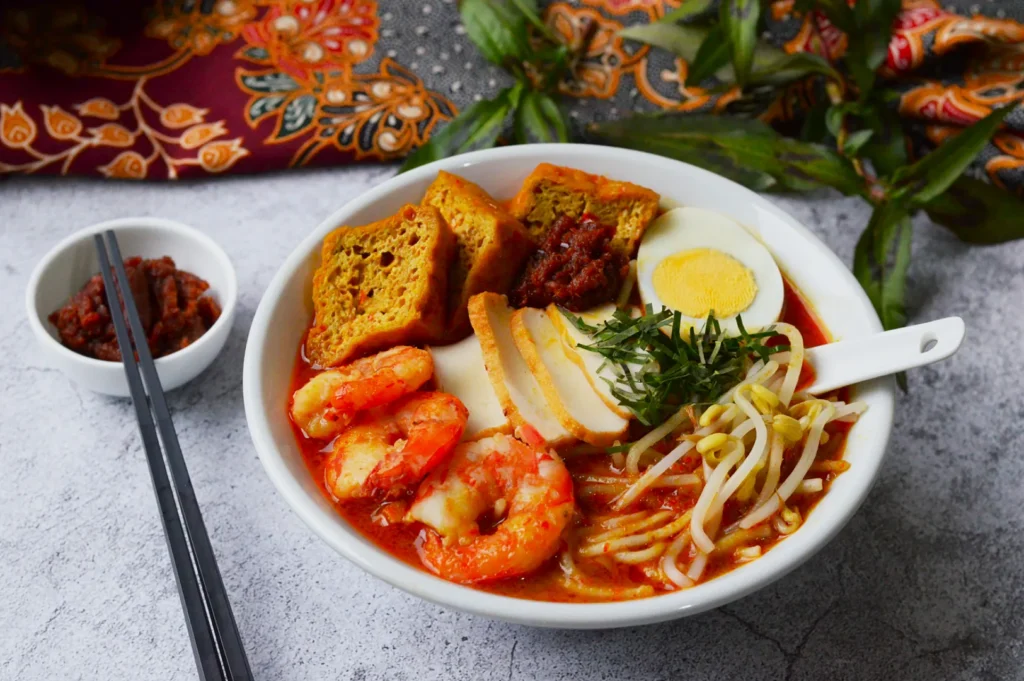 A colorful bowl of shrimp laksa is garnished with boiled egg, tofu, herbs, and bean sprouts, accompanied by chopsticks and sambal on the side.