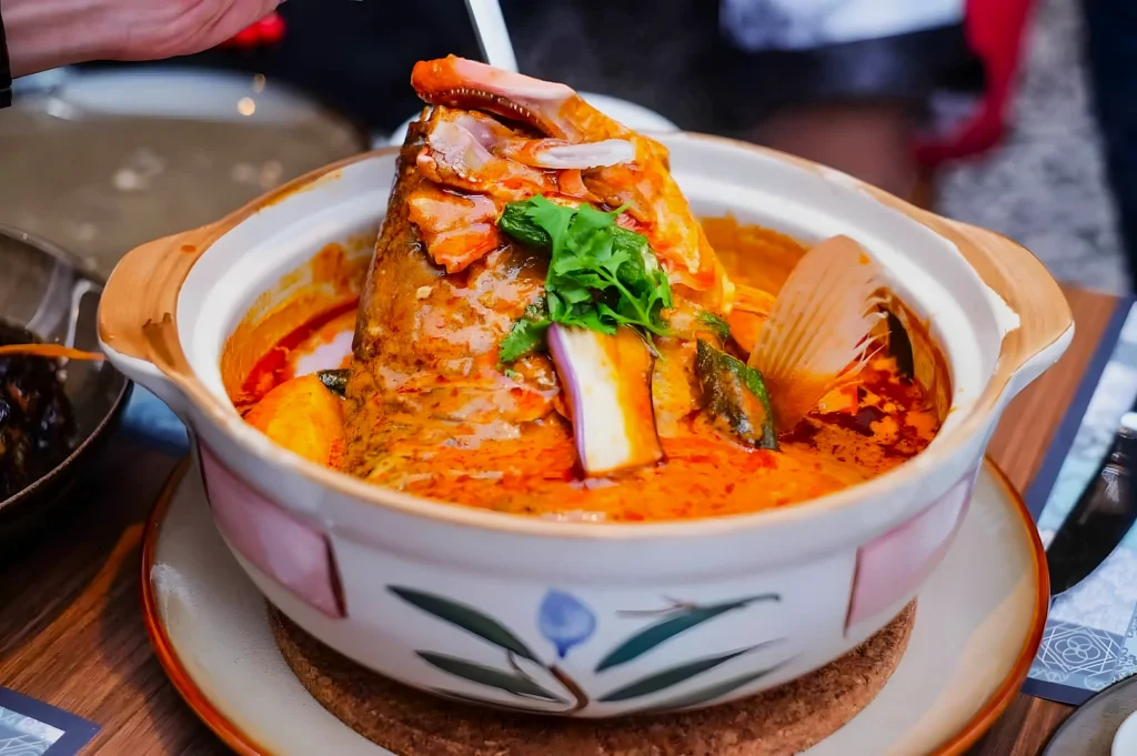A vibrant, spicy Malaysian fish head curry in a decorative ceramic pot. Garnished with fresh cilantro, it exudes a rich, inviting aroma.