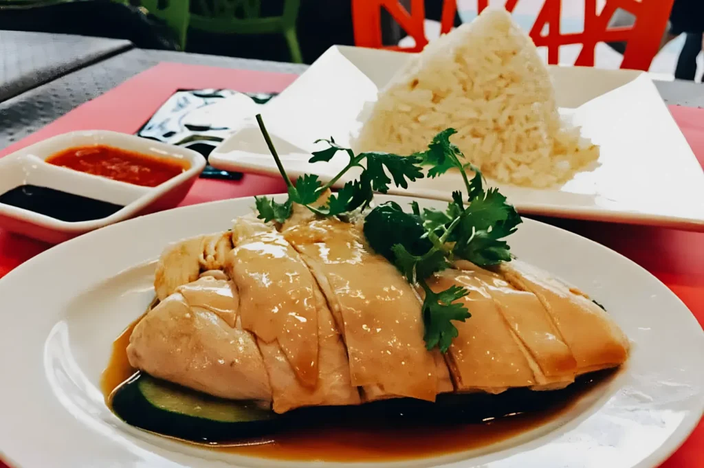 Poached chicken slices on soy sauce with cilantro garnish, served with a side of pyramid-shaped rice. Condiments include chili and soy sauces.