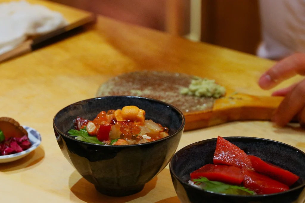 Two black bowls contain assorted sushi ingredients, including salmon and roe, on a wooden surface. A hand prepares wasabi in the background, conveying a fresh, culinary atmosphere.