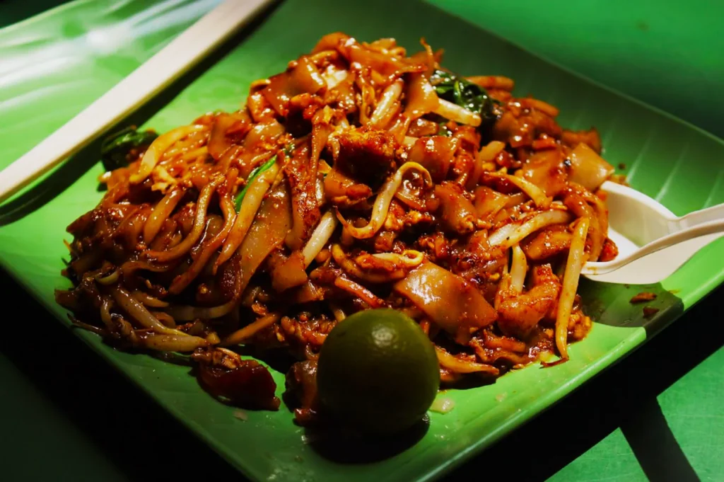 A plate of stir-fried noodles with vegetables, bean sprouts, and meat sits on a green leaf. A lime wedge and white utensils are beside the dish.