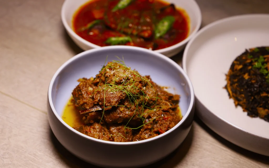 A bowl of spicy beef rendang garnished with herbs, surrounded by rich curry and a vegetable dish. The presentation is appetizing and vibrant.