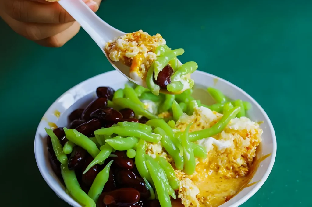 A hand holds a spoonful of cendol, featuring green jelly noodles, red beans, and crushed ice, from a white bowl. The dessert is vibrant against a green background.