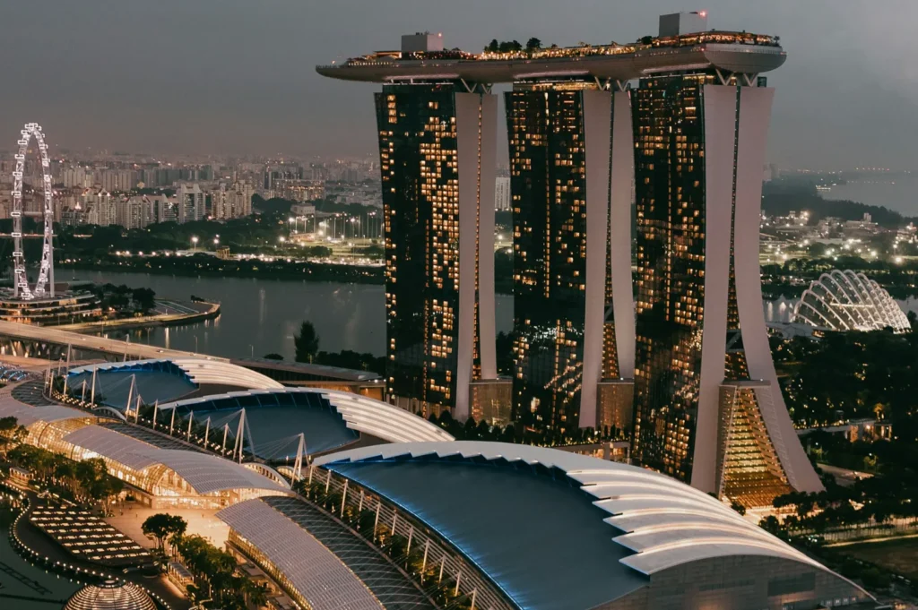 Aerial view of Marina Bay Sands in Singapore at dusk, featuring three illuminated towers topped by a sky park, with a cityscape and Ferris wheel in the background.