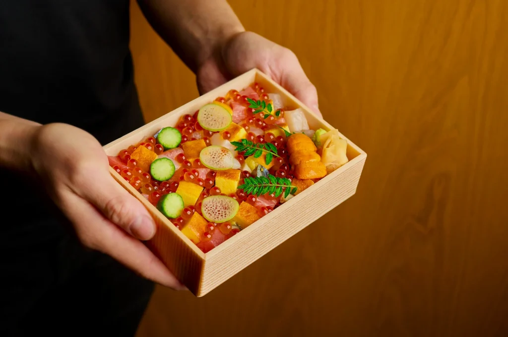 Hands hold a wooden box with colorful sushi, featuring salmon roe, sea urchin, cucumber slices, radish, and garnishes, against a wood background.