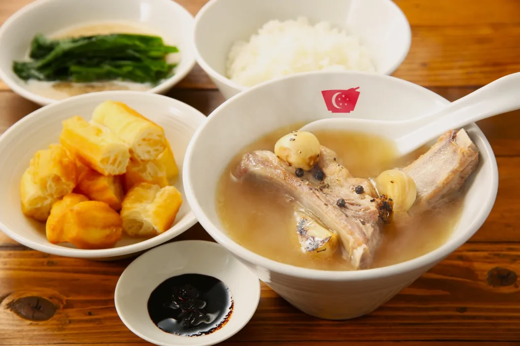 A bowl of bak kut teh with tender pork ribs, garlic, and peppercorns in broth, surrounded by rice, crispy breadsticks, greens, and soy sauce. Cozy and inviting.