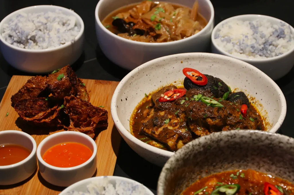 A variety of dishes featuring spiced curry with meat and vegetables garnished with red chili, steamed rice, crispy fried slices, and dipping sauces on a table.