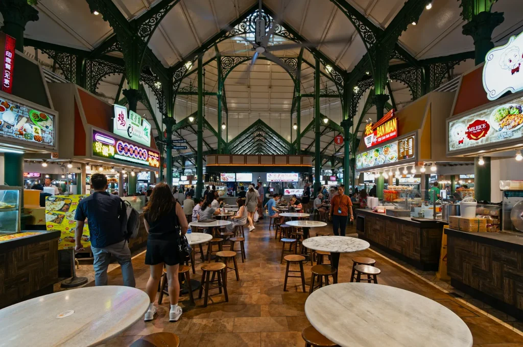 A spacious food hall with ornate green metal beams and a high ceiling. People browse diverse food stalls lined on both sides, creating a lively, bustling atmosphere.