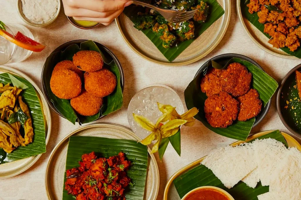 An assortment of vibrant South Asian dishes on a beige table, served on banana leaves. A hand reaches to take food, evoking a lively, communal meal.