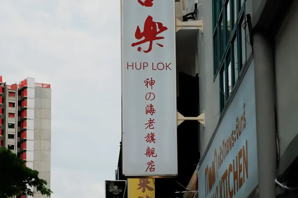 A vertical sign for "Hup Lok" with red Chinese characters is mounted on a building. Nearby, there's another building with a sign that says "Kitchen." A high-rise is visible in the distance, beneath a partly cloudy sky.