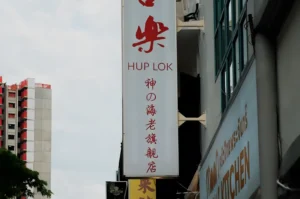 A vertical sign for "Hup Lok" with red Chinese characters is mounted on a building. Nearby, there's another building with a sign that says "Kitchen." A high-rise is visible in the distance, beneath a partly cloudy sky.