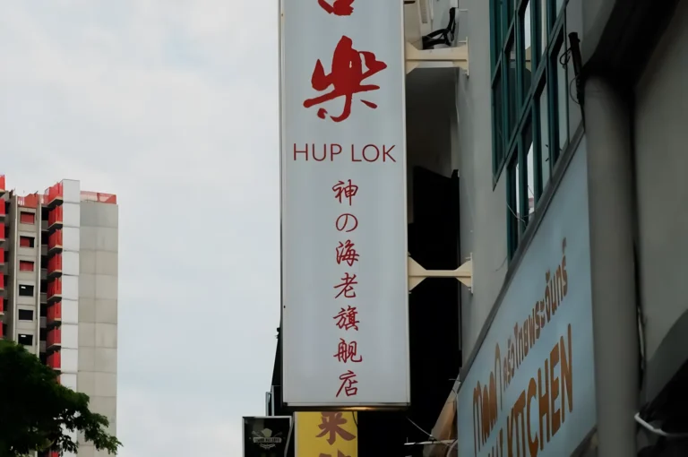 A vertical sign for "Hup Lok" with red Chinese characters is mounted on a building. Nearby, there's another building with a sign that says "Kitchen." A high-rise is visible in the distance, beneath a partly cloudy sky.