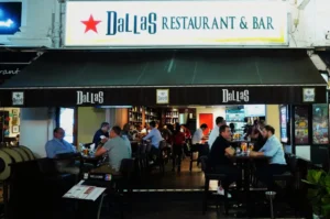 Outdoor seating at "Dallas Restaurant & Bar" with patrons enjoying drinks and conversation under a black awning in an inviting, lively atmosphere