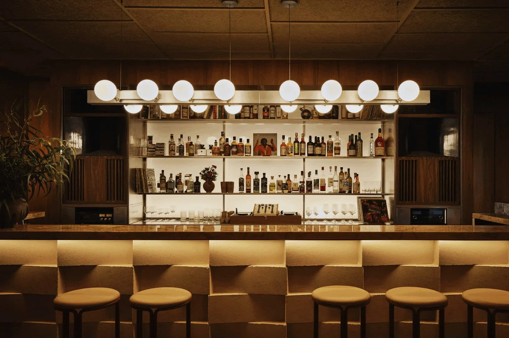 Dimly lit bar with a warm, inviting atmosphere. Modern globe lights hang above a stocked liquor shelf, while stools line the textured counter.