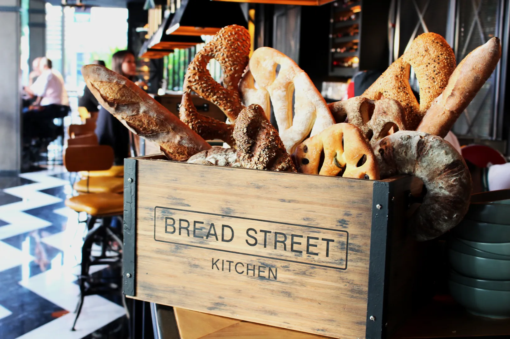 A rustic wooden box labeled "Bread Street Kitchen" holds an assortment of artisan bread, including baguettes and pretzels, in a chic restaurant setting.