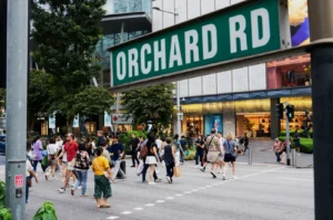 A bustling crosswalk on Orchard Road with people walking in various directions. A green street sign in the foreground adds context to the lively urban scene.