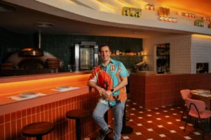 Chef Antonio Miscellaneo in a colorful shirt leans on a counter in a stylish pizzeria. The room features orange tiles, a pizza oven, and warm lighting, creating a cozy vibe