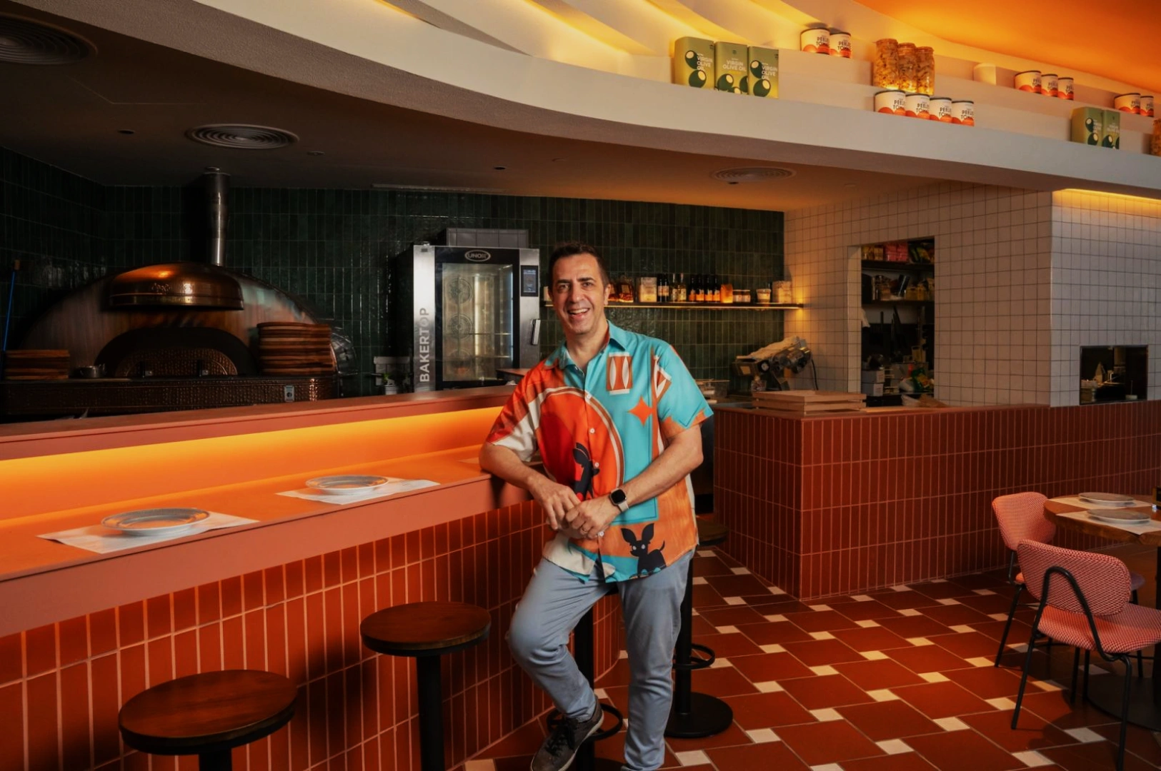 Chef Antonio Miscellaneo in a colorful shirt leans on a counter in a stylish pizzeria. The room features orange tiles, a pizza oven, and warm lighting, creating a cozy vibe