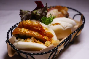 Two steamed bao buns filled with crispy chicken, lettuce, and herbs sit in a black wire basket, creating an appetizing and savory scene.