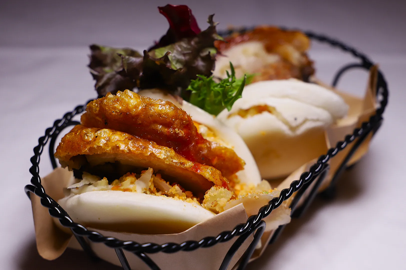 Two steamed bao buns filled with crispy chicken, lettuce, and herbs sit in a black wire basket, creating an appetizing and savory scene.