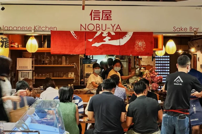 Japanese kitchen and sake bar with a lively crowd seated at the counter. Two staff members in masks interact with patrons beneath red banners. Warm, inviting atmosphere.