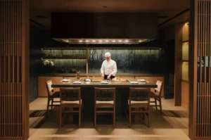 A chef in a white uniform stands at a modern kitchen island, preparing food. The room is warmly lit with wooden decor, giving a calm, inviting atmosphere.
